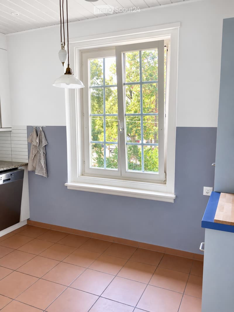 Modern kitchen with tiled floor, blue and white walls, large window with white frame, pendant light, and a dishwasher.