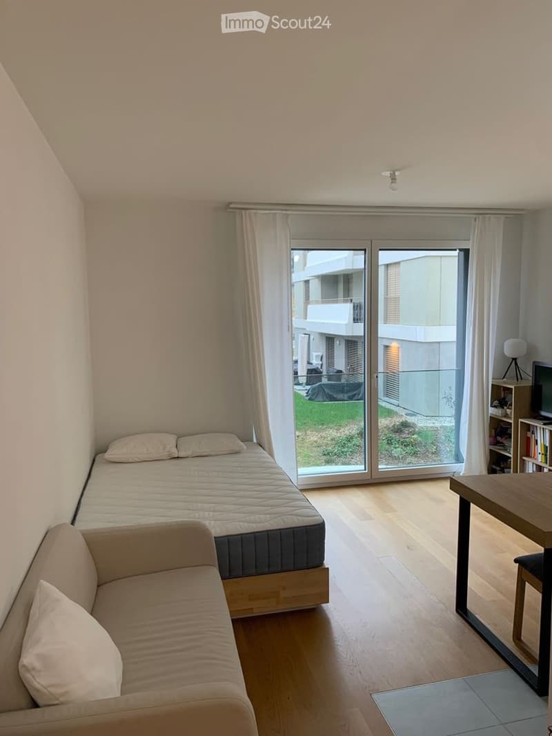 A modern, compact living space with a bed, sofa, small table, and bookshelf, featuring large glass doors leading to a balcony.