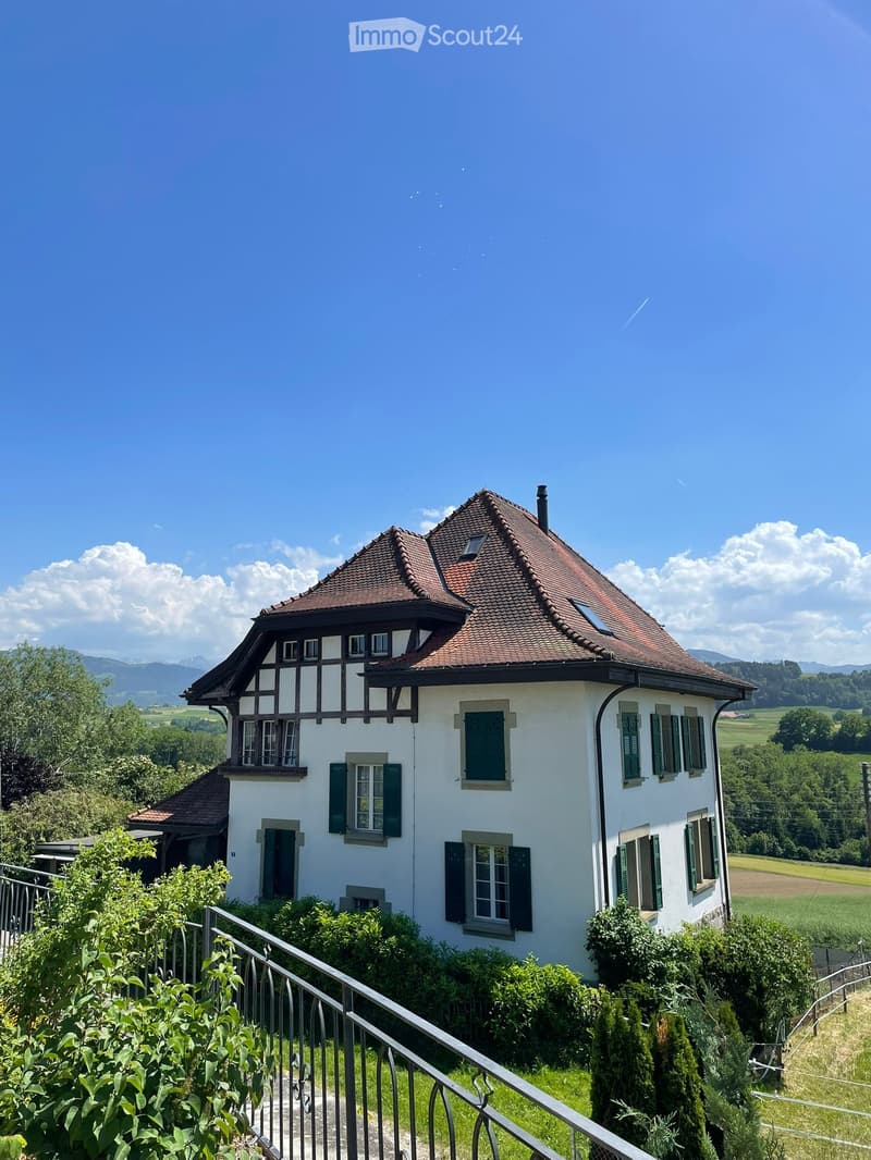 A two-story white house with a red-tiled roof, green shutters, and a balcony overlooking a scenic valley with mountains in the background.