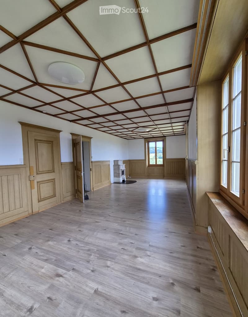 Empty hallway with wooden flooring, white walls, and a wooden ceiling with exposed beams. There are doors and windows along the walls, and a small white appliance is visible in the background.