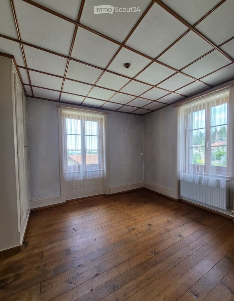 Empty room with wooden flooring, white walls, and large windows with sheer curtains. The ceiling features a geometric pattern with wooden beams and recessed lighting.