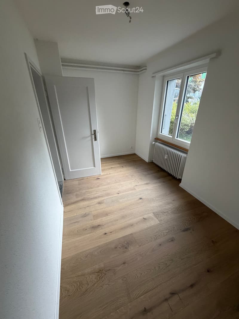 Empty room with light wooden flooring, white walls, a window with a radiator, and a closed door.