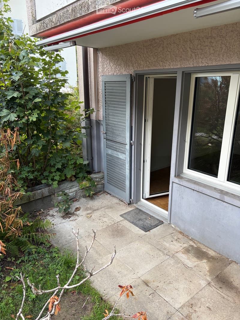 Concrete patio area adjacent to a building with a covered entrance, greenery, and a window with shutters.