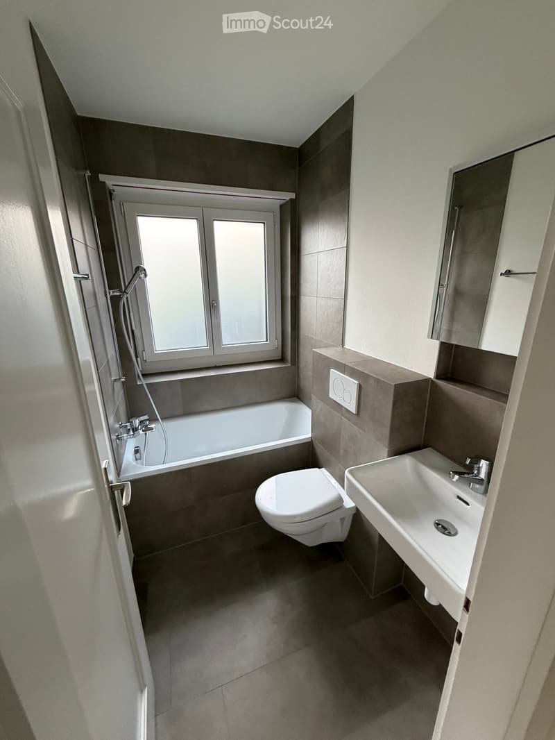 Modern bathroom with a bathtub, toilet, sink, and mirror. Features a window above the bathtub and a sleek, contemporary design with grey tiles and white fixtures.