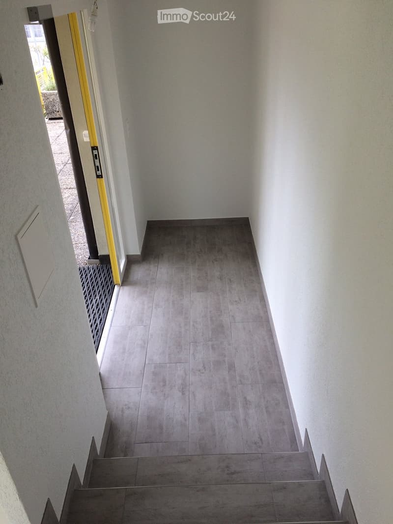 A clean, modern hallway with grey tiled flooring and white walls, featuring a staircase leading upwards and an open doorway to the left.