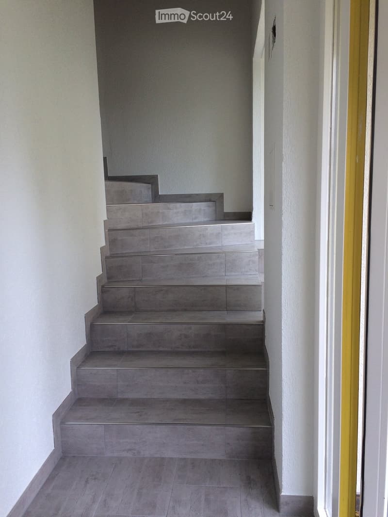A modern staircase with grey tiled steps and landings, leading upwards. The walls are painted white, and the floor is tiled in a matching grey tone. A golden door frame is visible on the right side.