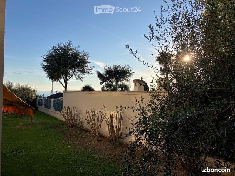Outdoor view of a house with a wall on the side, greenery in the area, including trees and shrubs, and a clear blue sky in the background. There is a grassy area with some plants on the left side of the wall. In the distance, there is a view of the sea. A