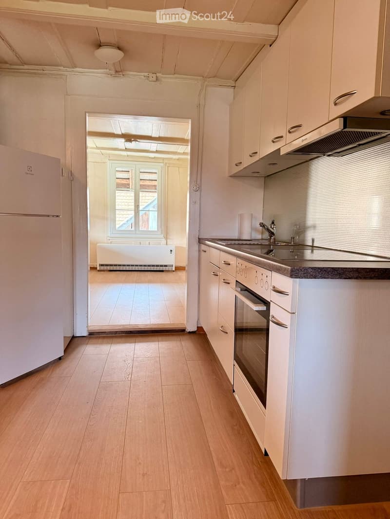 Modern kitchen with white cabinets, stainless steel appliances, and wooden flooring. An open doorway leads to a bright living area with large windows and a radiator.