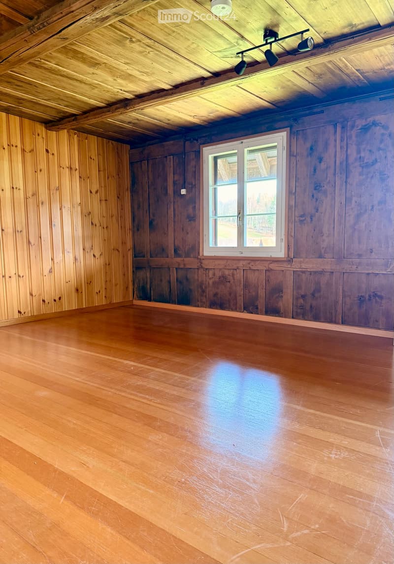 Empty room with wooden walls, wooden floor, and a single window with white frame. Exposed wooden ceiling with track lighting.
