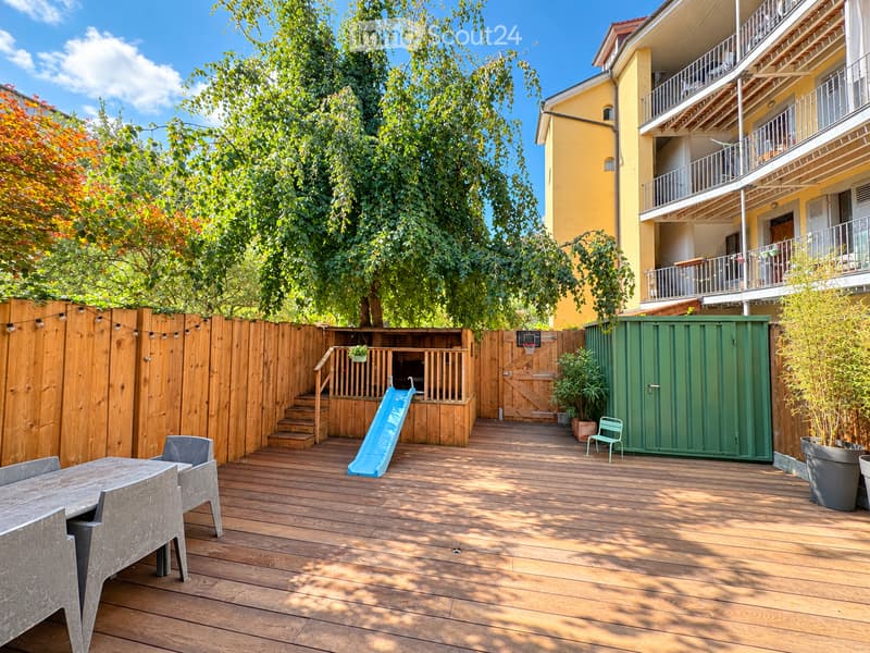 Wooden deck with a table, chairs, slide, small stool, and a basketball hoop, surrounded by a wooden fence and trees.
