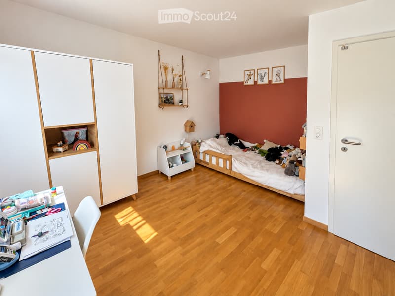 A well-lit children's bedroom with a wooden bed, white wardrobe, shelves, and a small table with toys and books.
