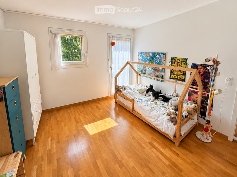 Bright, modern bedroom with wooden flooring, a lofted bed with stuffed animals, a wardrobe, and colorful posters on the wall.