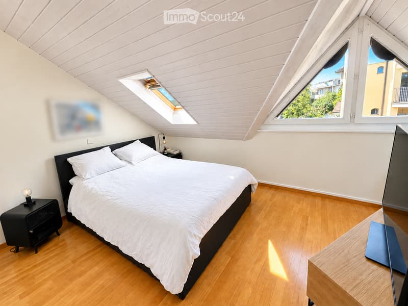 Bright attic bedroom with wooden floors, white walls, and a slanted wooden ceiling. Features a large bed with white bedding, a nightstand with a lamp, and a modern screen on a wooden stand. Large windows and a skylight provide natural light.