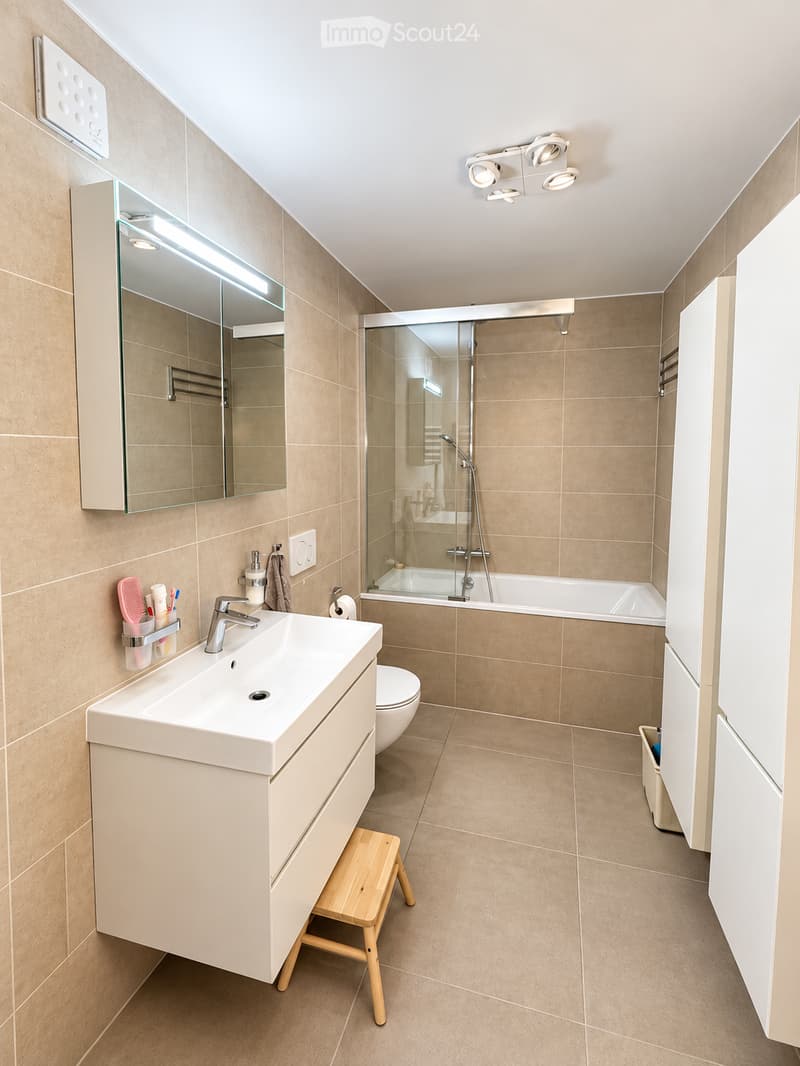Modern bathroom with beige tiles, a white sink with a modern faucet, a large mirror with built-in lighting, a toilet, a bathtub with a glass shower enclosure, and a small wooden stool.