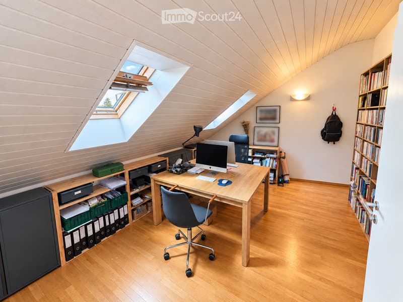 Bright, modern attic office with wooden floors, slanted ceilings, and large skylights. Features a wooden desk with a computer, chair, and storage shelves, along with bookshelves and a hanging backpack.