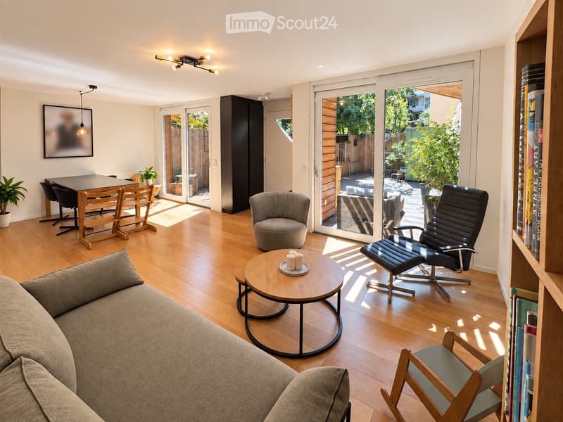 Modern living room with hardwood floors, large windows, and sliding glass doors leading to a patio. Features a grey sofa, round coffee table, black leather chair, dining table with chairs, bookshelf, and potted plants.