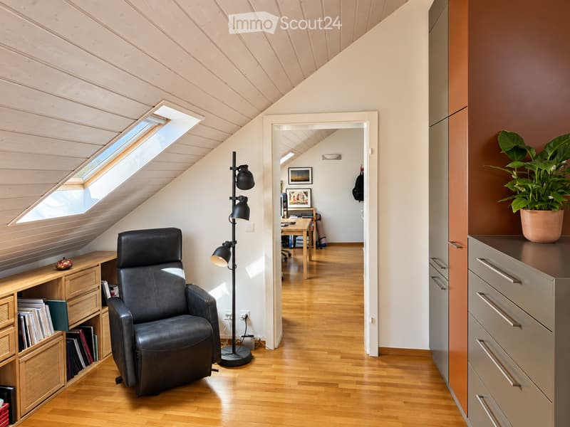 Modern, well-lit room with wooden flooring, slanted wooden ceiling, skylight window, black leather chair, floor lamp, wooden cabinet with books, and a potted plant on a grey cabinet.
