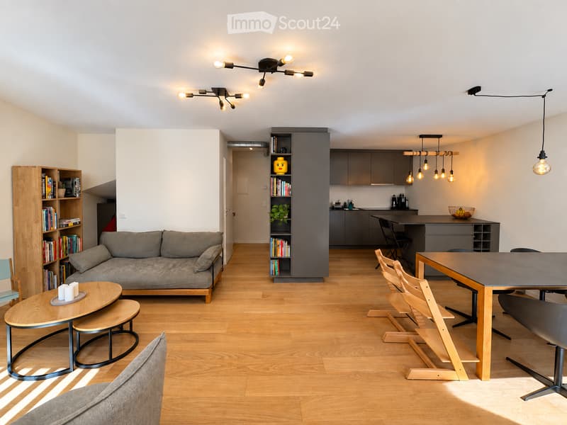 Modern open-plan living and dining area with light wooden flooring, grey sofa, coffee table, dining table with chairs, bookshelf, and kitchen area with cabinets and pendant lights.