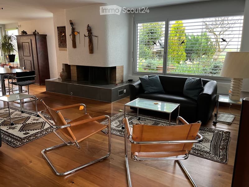 Modern living room with hardwood flooring, large windows, a black leather sofa, glass coffee table, two leather chairs, a fireplace, and decorative items including a cabinet and vases.