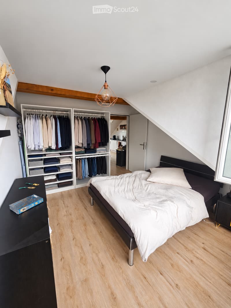 Modern bedroom with wooden flooring, a large bed with white bedding, a built-in wardrobe with hanging clothes and folded linens, a nightstand with a lamp, and a small drawer unit.