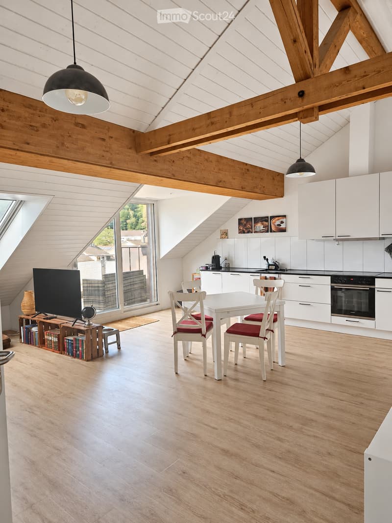 Open-plan living and dining area with exposed wooden beams, white cabinets, a dining table with chairs, a television, and a small bookshelf with books and a vase.