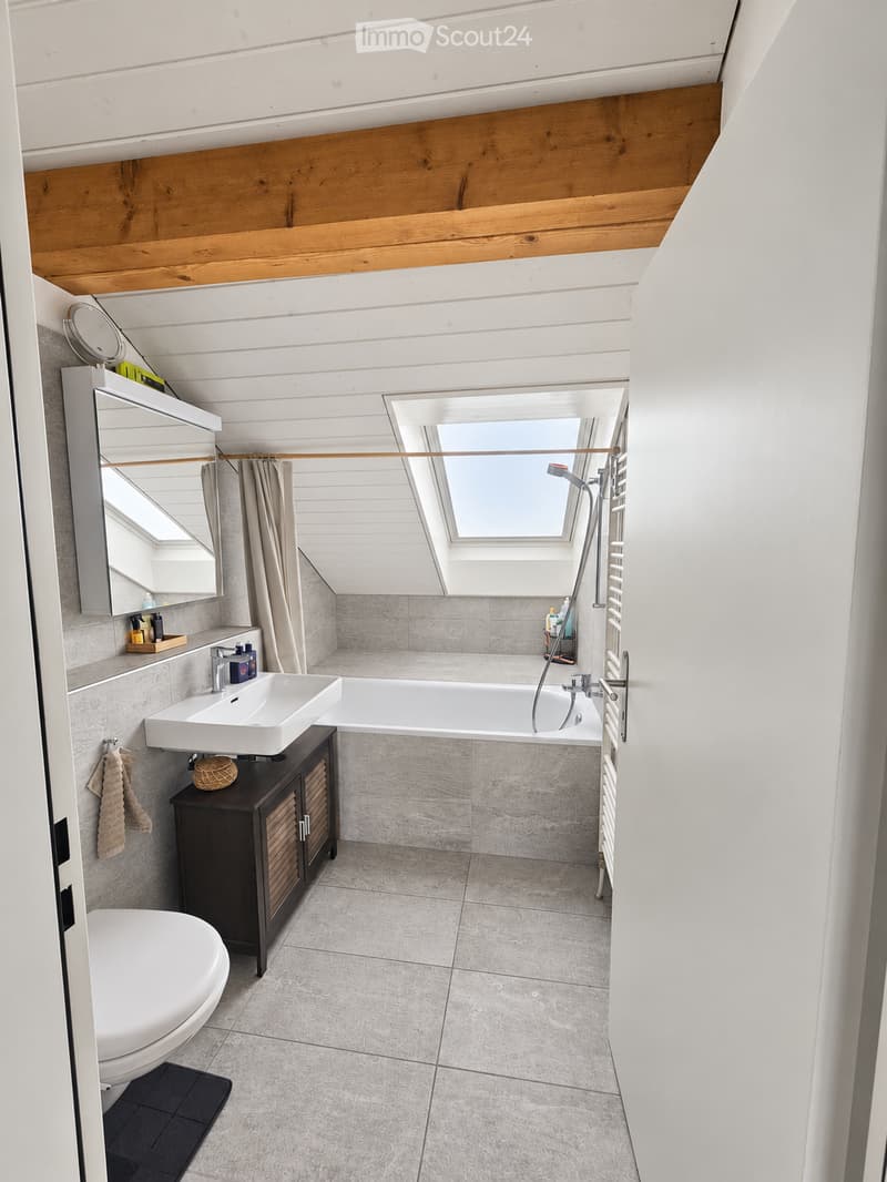 Modern bathroom with a white sink, toilet, bathtub, and a large mirror. The room features wooden slatted ceilings and large windows, with a dark wooden cabinet under the sink and a towel hanging on the wall.