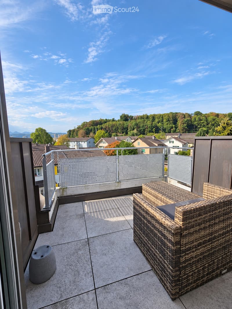 Balcony with a wicker chair, a grey stool, and a metal railing, overlooking a suburban area with houses and trees.