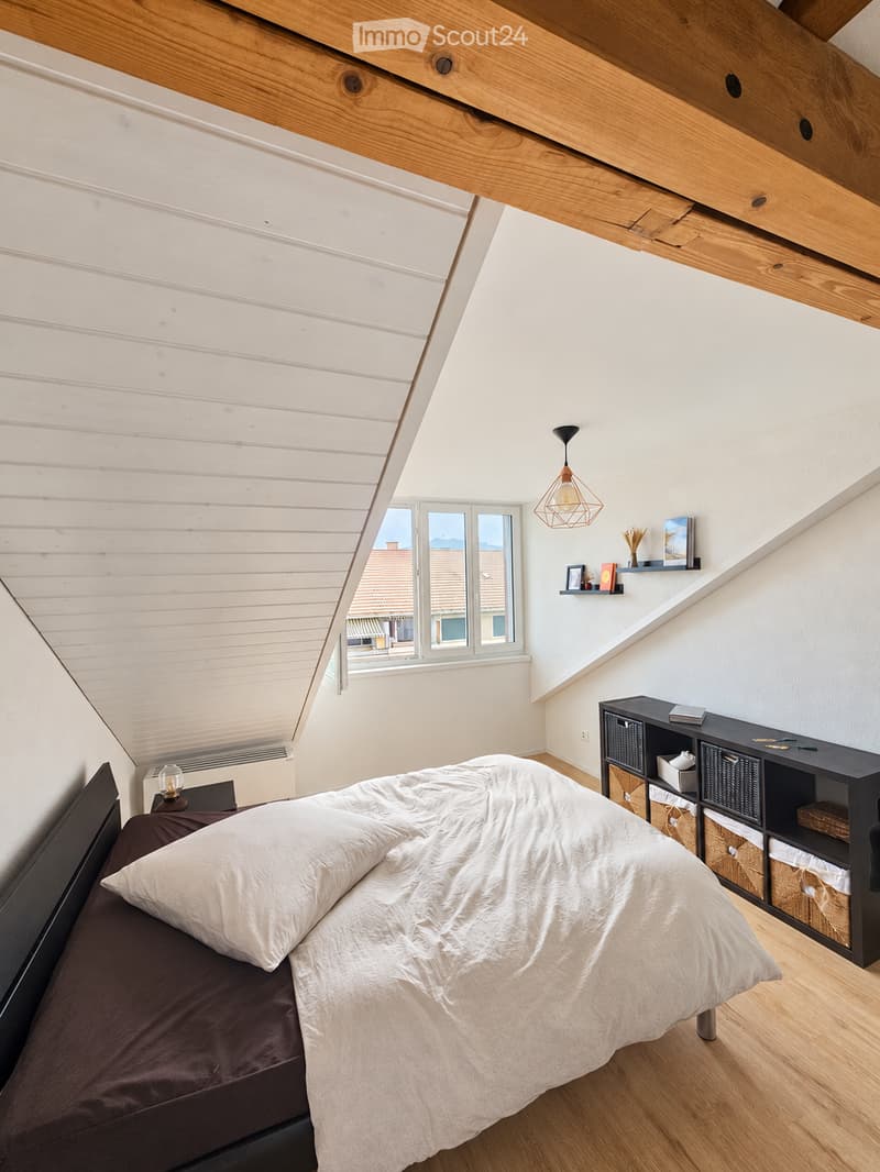 Cozy attic bedroom with wooden slanted ceilings, exposed beams, and large windows offering a view. Features a dark wooden bed with white bedding, a nightstand with a lamp, a black shelving unit with baskets, and a small shelf with books and decorative ite