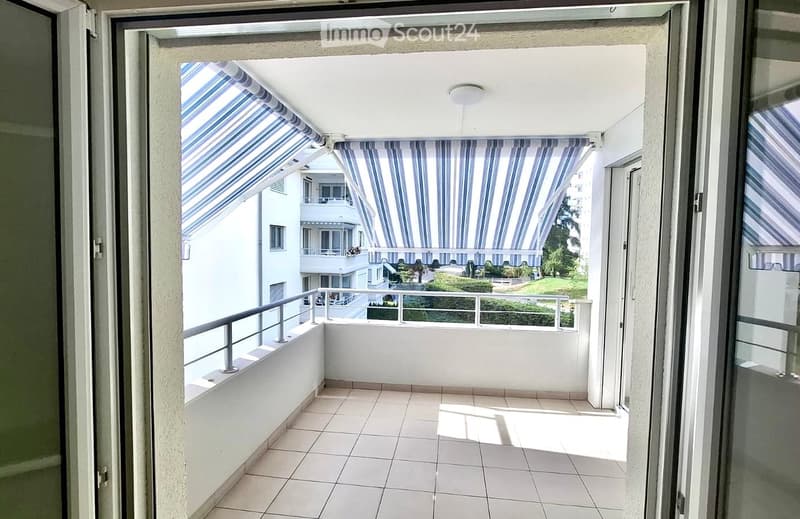 Balcony with tiled floor, white walls, metal railing, and striped awnings providing shade. The view shows neighboring buildings and greenery.