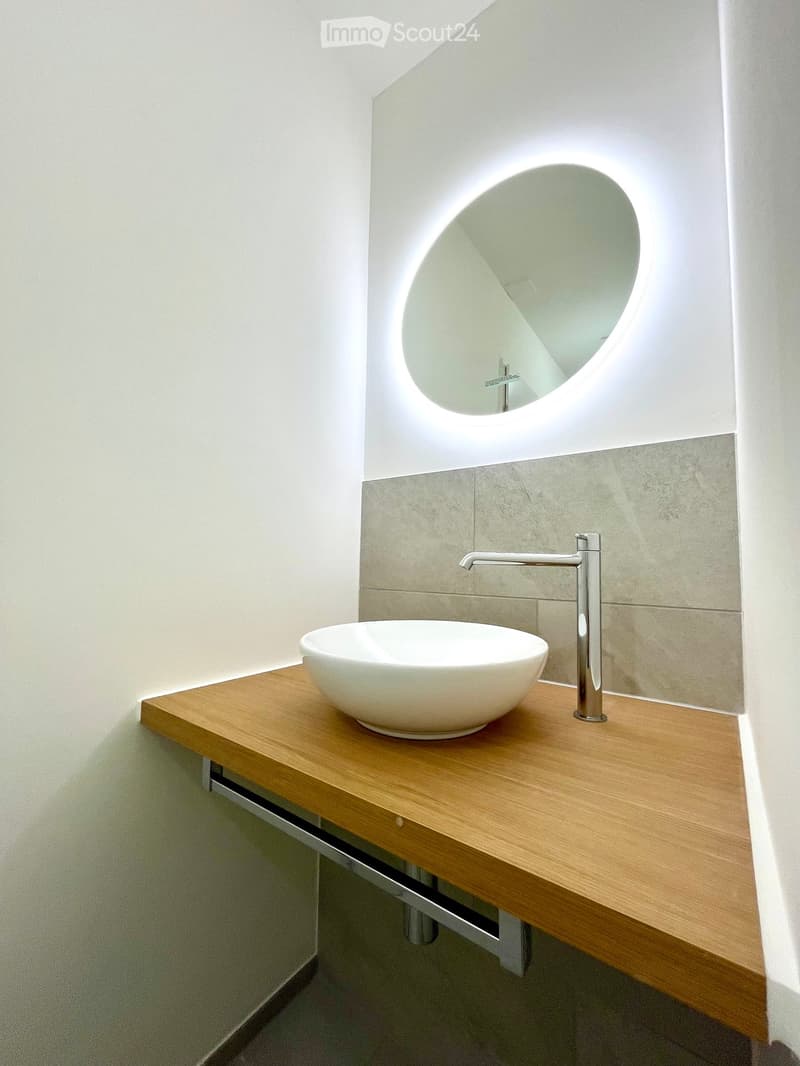 Modern bathroom with a wooden countertop, white sink bowl, sleek chrome faucet, and a round mirror with a built-in light fixture.