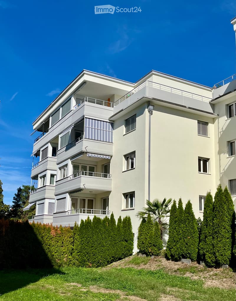 Modern two-story white building with multiple balconies, large windows, and neatly trimmed hedges in the front yard.