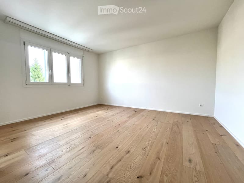 Empty room with light wooden flooring and white walls, featuring a large window with white frames and a visible electrical outlet on the wall.