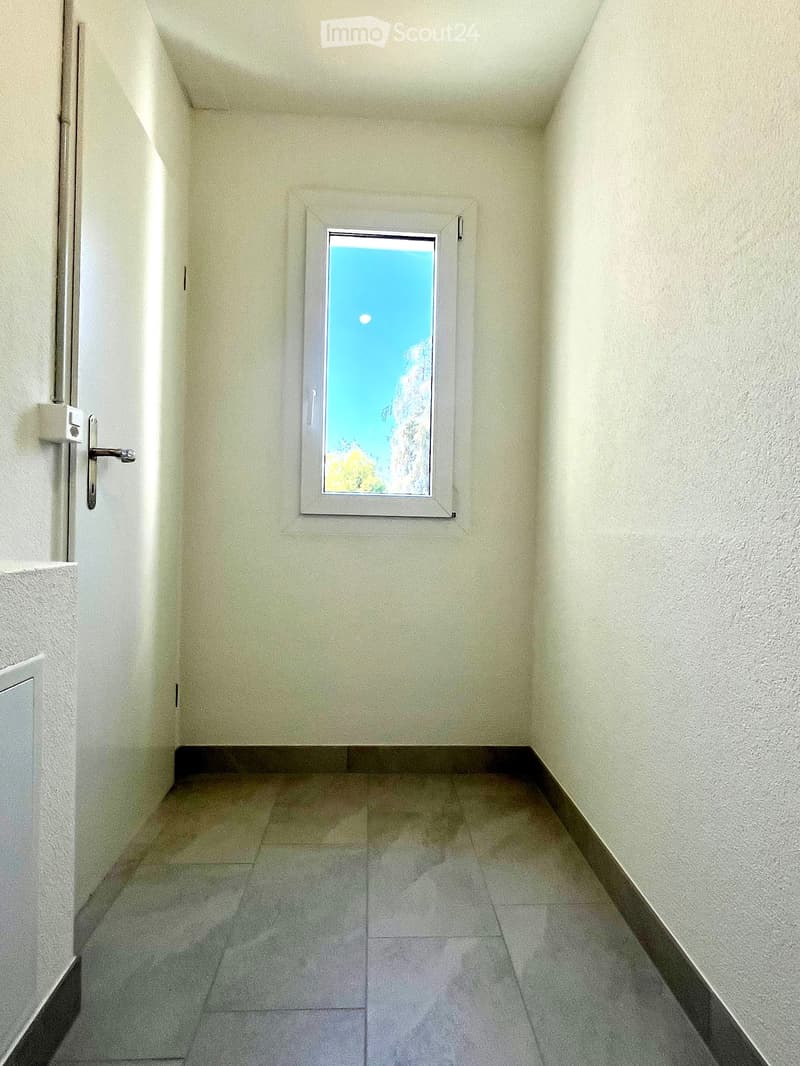 Empty hallway with tiled floor, light-colored walls, and a single window showing a view of the sky and greenery.