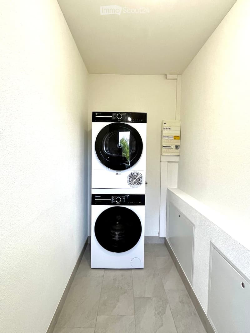 A small, clean room with a white washing machine and dryer stacked vertically, a control panel, and a tiled floor.