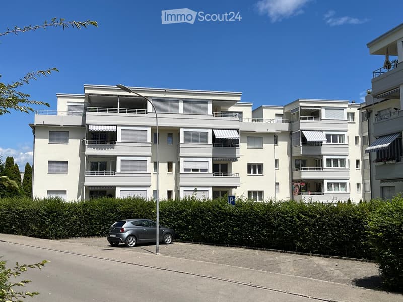 Modern white multi-story apartment building with balconies, awnings, and surrounding greenery.