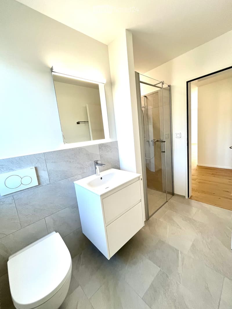 Modern bathroom with a white sink, toilet, large mirror, and glass-enclosed shower. Grey tiles on lower walls and light-colored flooring.
