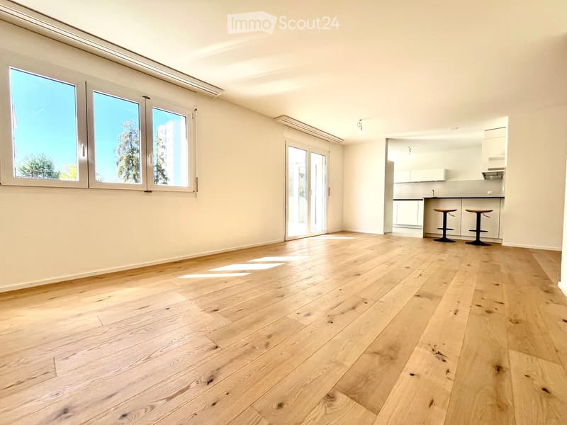 Bright, open living space with light wooden flooring, large windows, and a sliding glass door leading to a balcony. A small kitchen area is visible with two stools at the counter.