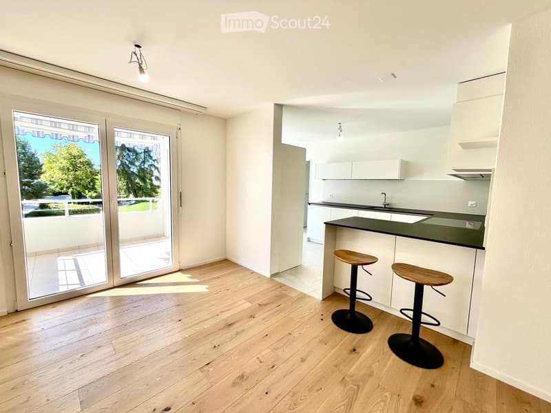 Modern kitchen with light wooden flooring, white cabinets, black countertop, and two wooden stools. Large sliding glass doors lead to a balcony with outdoor view.