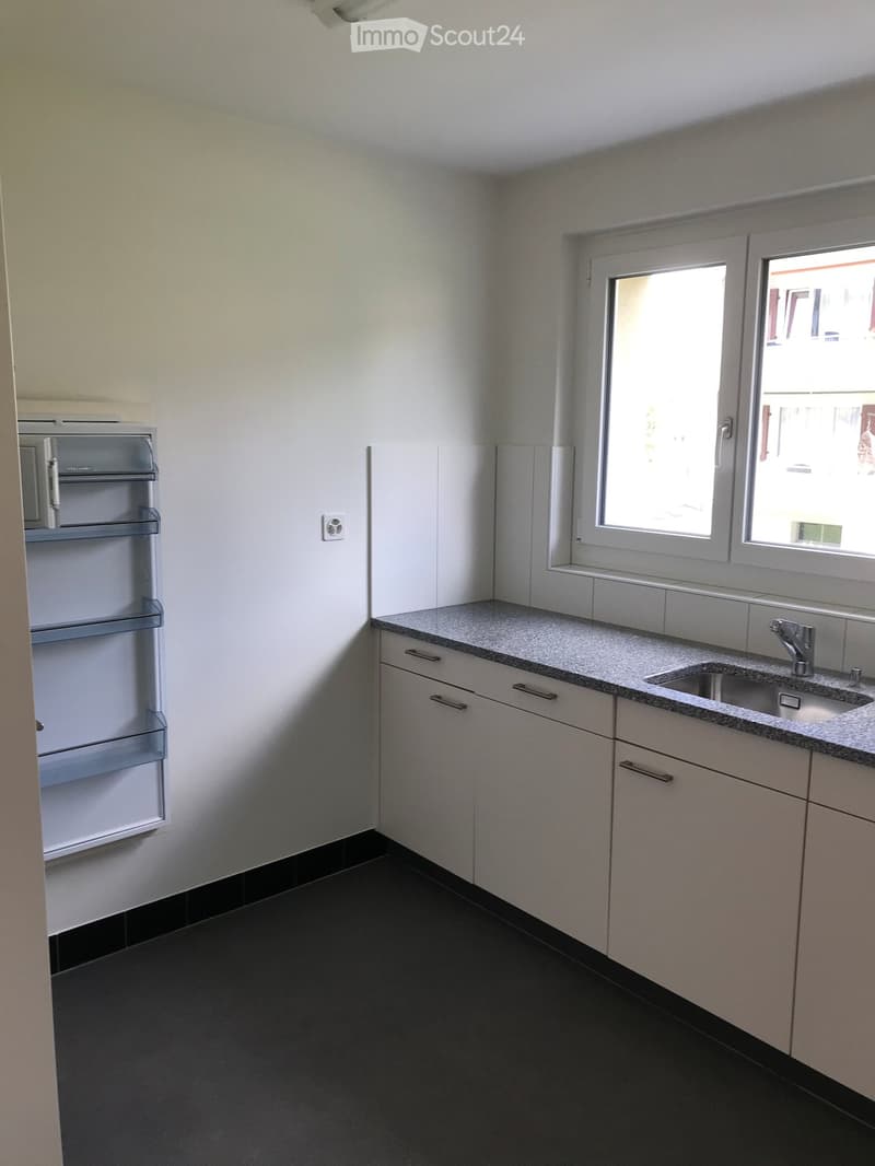 Modern kitchen with white cabinets, granite countertop, stainless steel sink, and a built-in refrigerator. The room features large windows and a dark tiled floor.