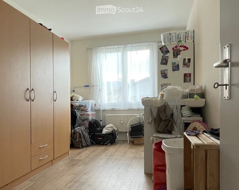 Wooden floor, white walls, wooden wardrobes, clothes on racks, white radiator, white washing machine, white window with white curtains, shelves with storage boxes