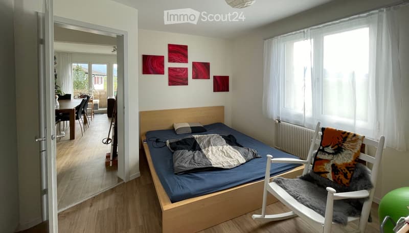 single bed, white wall, wooden floor, wooden headboard, small white chair, bedside table, radiator, large windows with white curtains