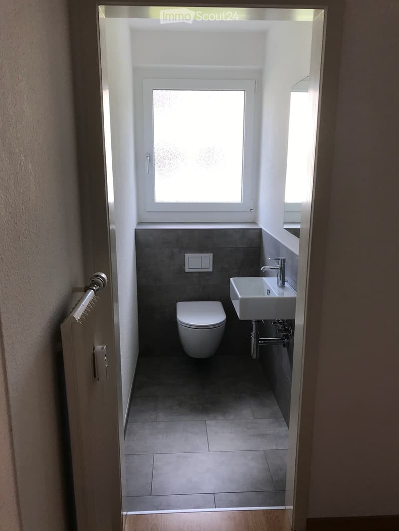 Modern bathroom with a toilet, sink, and faucet, featuring grey tiles and a frosted window above the sink.