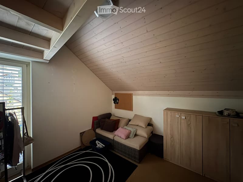 Cozy attic room with slanted wooden ceiling, large window with blinds, sofa with multiple pillows, wooden cabinet, small stool, and a patterned rug.