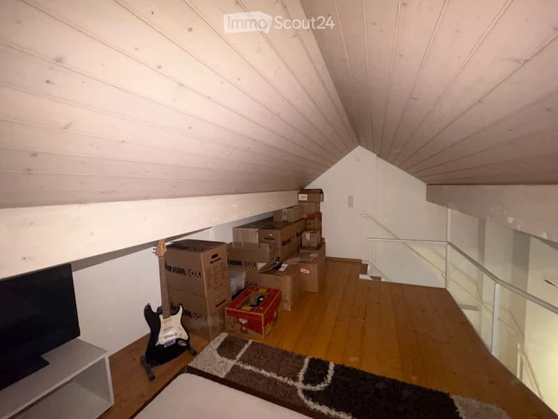 An attic room with wooden flooring, slanted white wooden ceiling, and several cardboard boxes stacked against the wall. A guitar is placed on a small table, and a screen is mounted on the wall. A small rug is on the floor.
