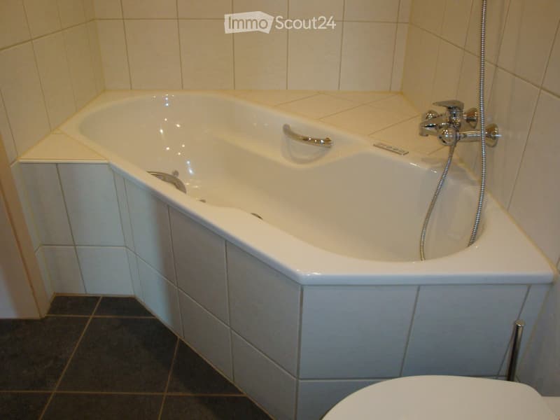 Bathroom with a white bathtub, chrome faucet, and a toilet. The walls are tiled with light-colored tiles, and the floor has dark tiles.