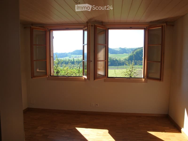 Empty room with wooden flooring, light-colored walls, and large windows offering a scenic view of a green landscape.