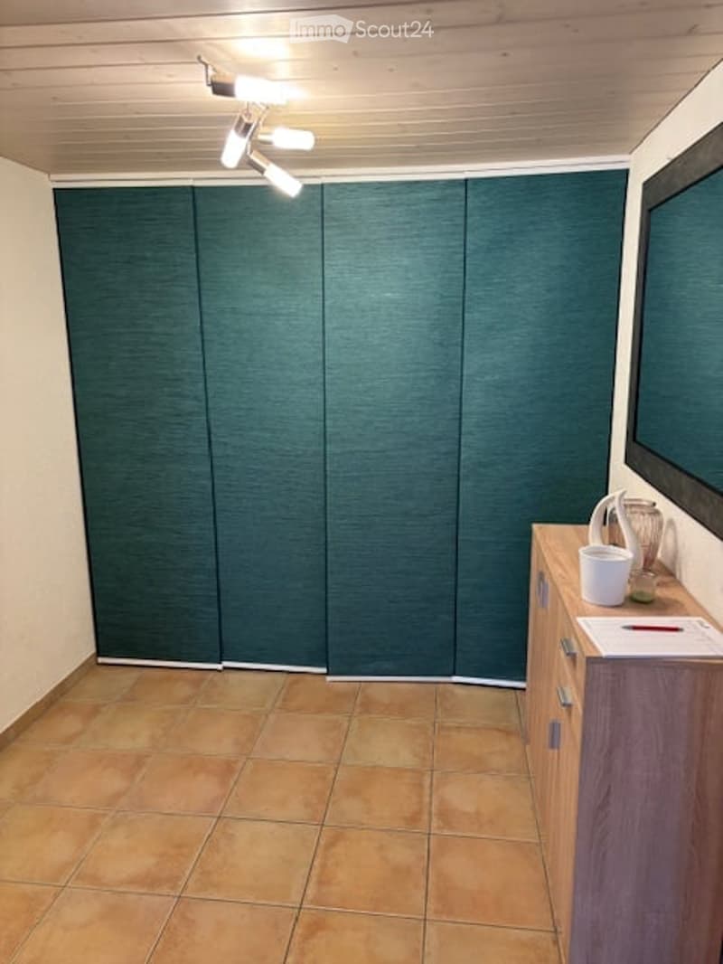 A small, unfurnished room with tiled flooring and a wooden cabinet. A large green screen or partition is mounted on the wall, and a mirror is positioned on the right side.