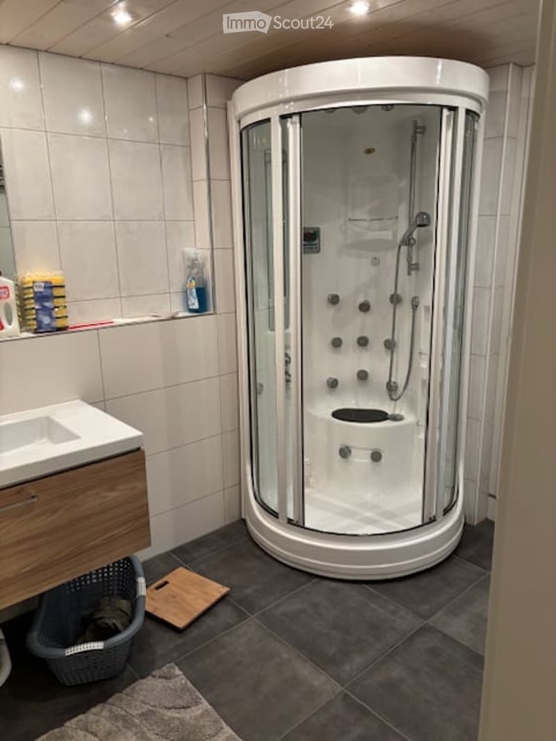 Modern bathroom with a round glass-enclosed shower stall, a white sink with a wooden cabinet, and a tiled floor. Cleaning supplies and a laundry basket are visible on the counter and floor.