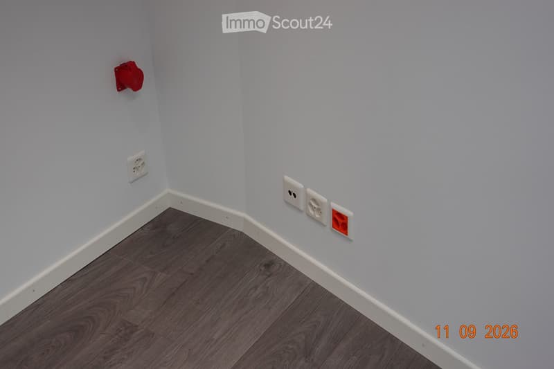 Empty room with light grey walls and dark wooden flooring, featuring electrical outlets and a red fire alarm box on the wall.
