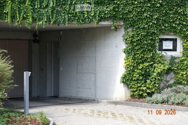 Modern concrete building entrance with ivy-covered walls, small garden area, and signage reading 'Studio / Büro / Hobbyraum'.